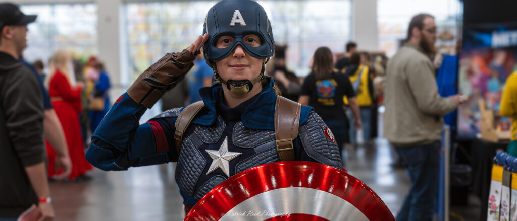 "A female cosplayer dressed as Captain America giving a salute, wearing a patriotic costume with a star-emblazoned chest and shield, embodying a modern twist on the classic superhero."